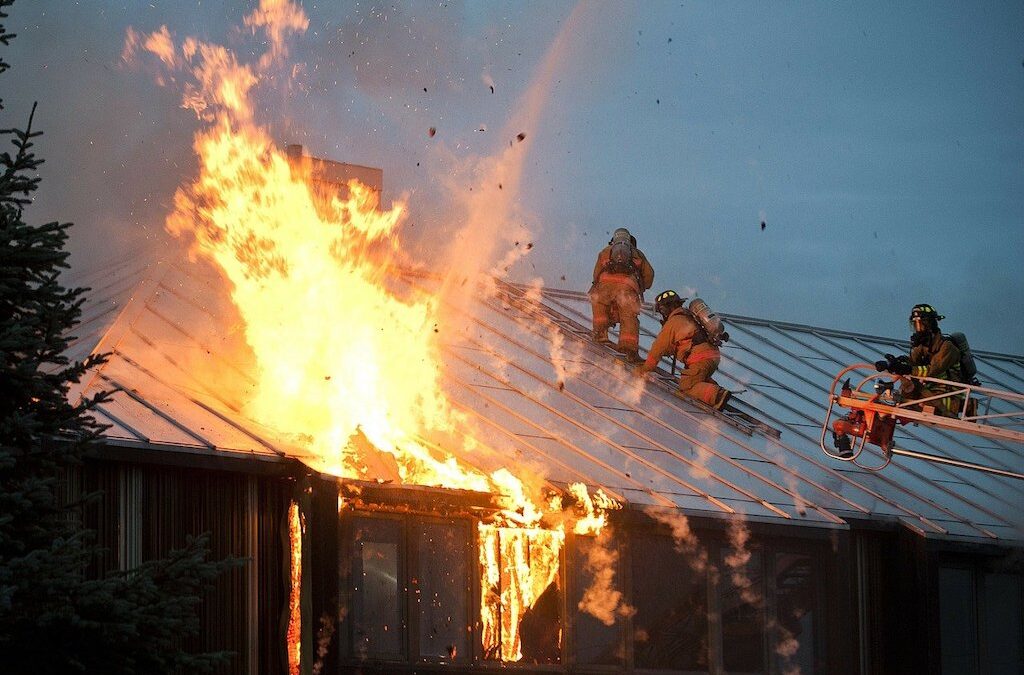 Photo of firefighters battling a house fire with flames shooting above a metal roof